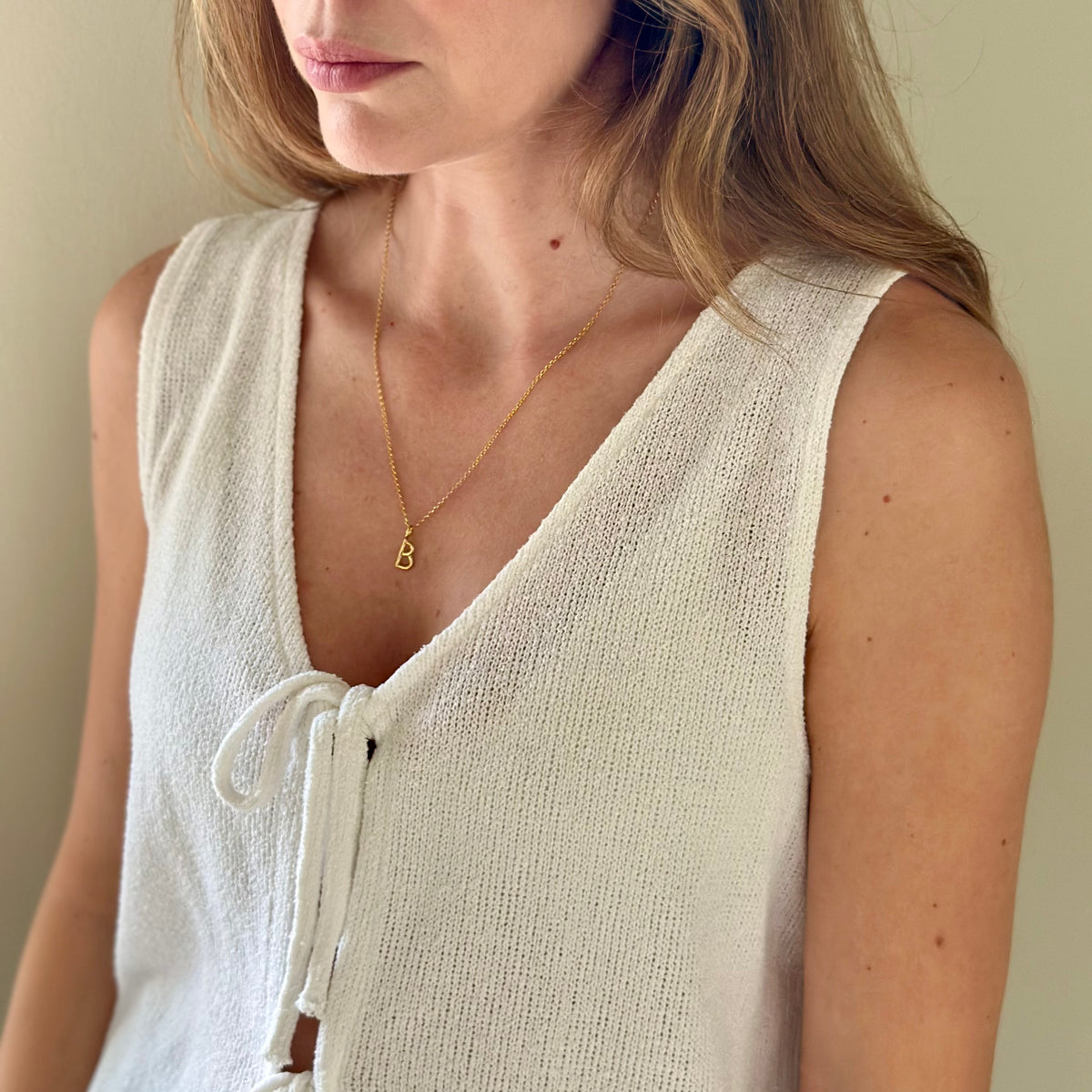 A close-up of a woman wearing a delicate gold chain necklace with a small letter 'B' pendant, paired with a cream-colored knit sleeveless top.