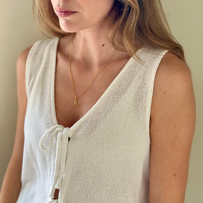 A close-up of a woman wearing a delicate gold chain necklace with a small letter 'B' pendant, paired with a cream-colored knit sleeveless top.