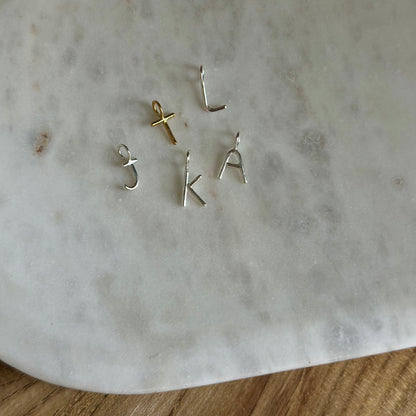 Five small letter and symbol charms, including three silver letters (J, K, A, T,L) arranged on a white marble surface.