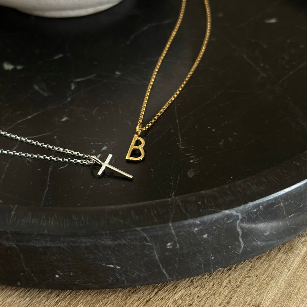 Two delicate initial necklaces displayed on a black marble tray. One necklace is gold with a box chain and a letter 'B' pendant, while the other is silver with a fine chain and a small 'T' pendant.